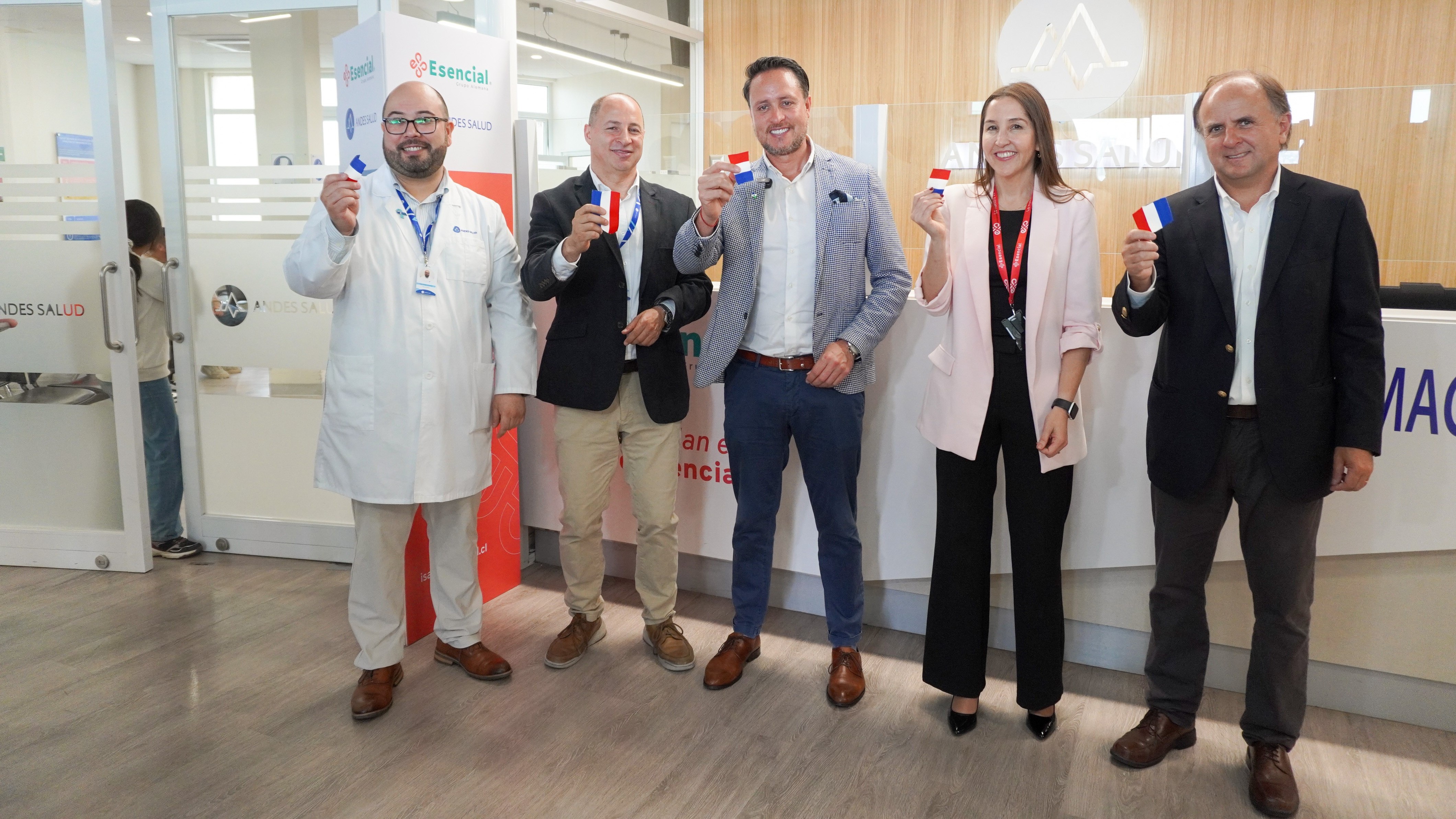 Grupo de 5 personas sonriendo a la cámara, todos sujetando una bandera pequeña en sus manos. Entre los miembros se encuentran personas tanto de Andes Salud como de Isapre Esencial.