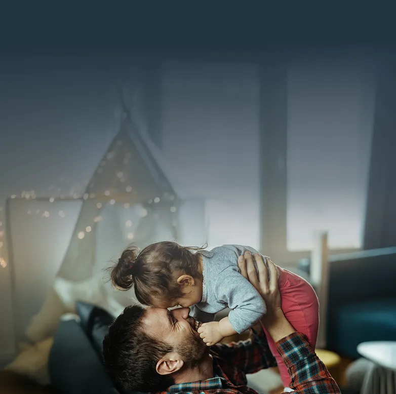 Fotografía de un padre levantando en brazos a su hija, ambos felices y sonriendo dentro de su casa. Al fondo se observa, desenfocado, un sillón azul y una carpa infantil con luces.