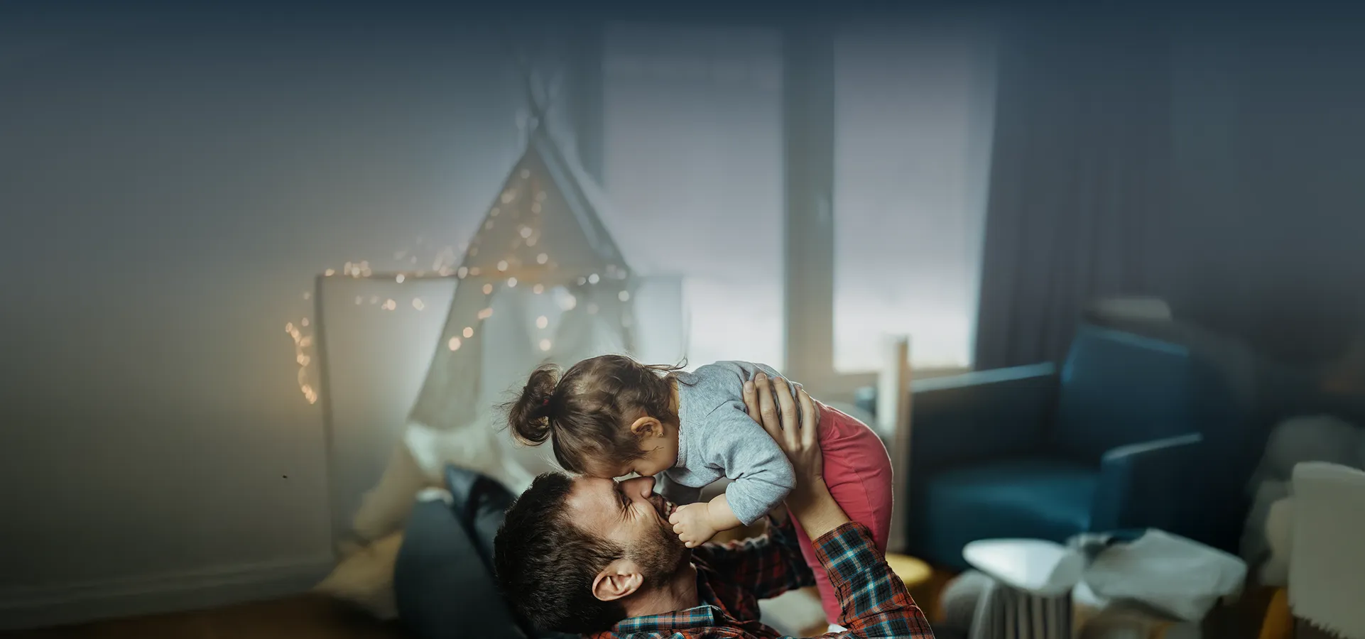 Fotografía de un padre levantando en brazos a su hija, ambos felices y sonriendo dentro de su casa. Al fondo se observa, desenfocado, un sillón azul y una carpa infantil con luces.