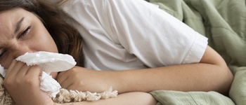 Mujer joven acostada en la cama y cubierta con una manta, con síntomas visibles de influenza, como fiebre y congestión nasal, usando un pañuelo.